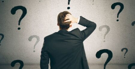 Homem de terno olhando para vários pontos de interrogação, simbolizando dúvidas e indecisão sobre escolhas importantes.